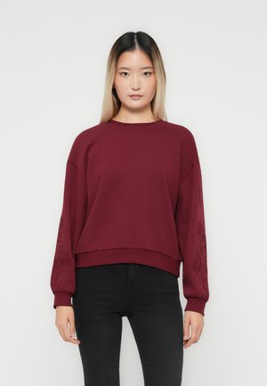 Ungdommelig kvinne med langt ombre-hår iført en burgunder genser med broderte ermer og svarte bukser, som står foran en ensfarget bakgrunn.