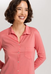Camicia a maniche lunghe color corallo in tessuto morbido e liscio, con chiusura a bottoni e una tasca sul petto. Caratterizzata da un colletto classico e una vestibilità aderente.