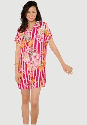 Lächelnde Frau, die ein kurzärmliges pink-weiß gestreiftes Kleid mit orange-pinkem Blumenmuster trägt, steht mit einem ausgestreckten Arm.