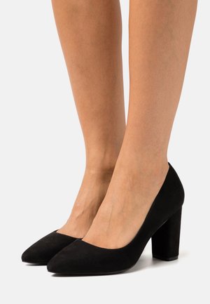 Tacones altos de ante negro con puntera puntiaguda y tacón grueso, con una textura suave y un diseño minimalista.