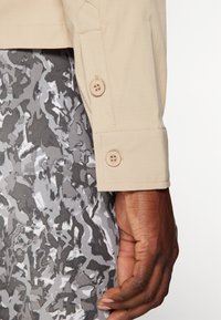 Camicia beige a maniche lunghe con polsini a bottoni, abbinata a pantaloni grigi multicolore con un motivo a strisce e design astratto testurizzato.
