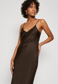 Robe nuisette en satin marron avec des bretelles fines et réglables, un décolleté en V profond et une silhouette élégante et fluide. La texture lisse renforce l'élégance.