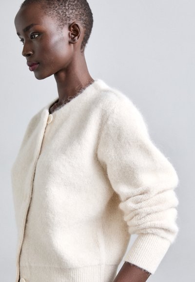 Cardigan spesso in crema realizzato in morbida lana strutturata. Presenta un collo rotondo, chiusura con bottoni sul davanti e polsini a coste per un tocco in più.