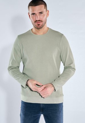 Mann mit kurzem Haar und Bart, trägt ein hellgrünes langärmeliges Hemd und blaue Jeans, steht vor einem einfarbigen hellen Hintergrund.