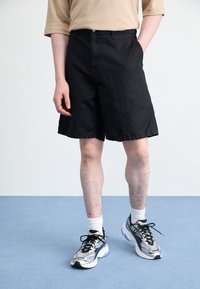 Schwarze Shorts mit lockerer Passform, aus strukturiertem Stoff. Kombiniert mit sportlichen Sneakern und weißen Socken, stehen auf einer hellblauen Fläche.