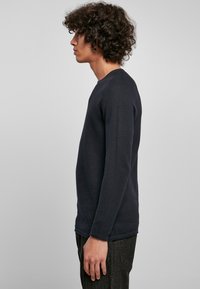 Tamsiai mėlyna ilgomis rankovėmis marškiniai iš tekstūruoto audinio, turintys apvalią iškirptę ir tiesų siluetą. Vilkima su tamsiomis kelnėmis, rodoma iš šono.