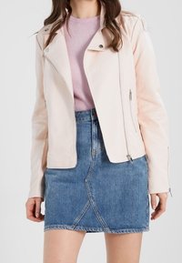 Helle rosa Kunstleder-Motorradjacke mit asymmetrischem Reißverschluss, zwei seitlichen Taschen, getragen über einem lavendelfarbenen Oberteil und einem Jeansrock.