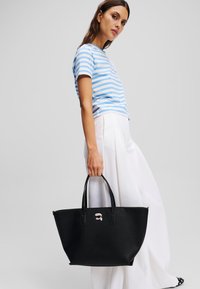 Zwarte textuur schoudertas met twee handvaten, voorzien van een grafische accent op de voorkant. Model draagt een blauw-wit gestreept shirt met witte broek.