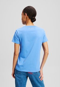 Kék pamut póló rövid ujjakkal, kerek nyakkivágással, és logó részlettel a hátsó részén. Kék farmernadrággal viselve.