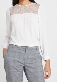 Blusa bianca a maniche lunghe con una yoke in pizzo trasparente, abbinata a pantaloni grigi a scacchi. La blusa presenta un scollo rotondo e una texture morbida.