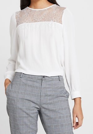 Blusa bianca a maniche lunghe con una yoke in pizzo trasparente, abbinata a pantaloni grigi a scacchi. La blusa presenta un scollo rotondo e una texture morbida.