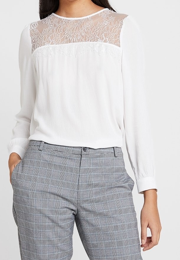 Weiße Bluse mit langen Ärmeln und transparenter Spitzenbesatz, kombiniert mit grauen Karohosen. Die Bluse hat einen Rundhals-Ausschnitt und eine weiche Textur.