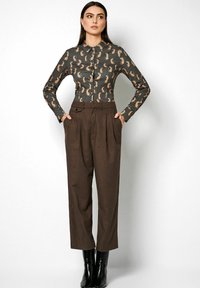 Long-sleeve blouse with black and tan pattern, paired with high-waisted brown trousers and black ankle boots, hands in pockets.
