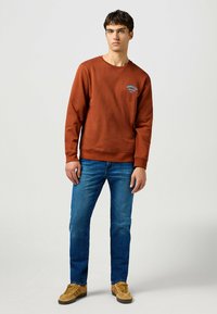 Felpa marrone con logo blu sul petto, abbinata a jeans blu e sneaker beige. Look casual e materiale in cotone.