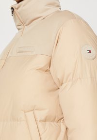 Beige péřová bunda s vysokým límcem, zapínáním na zip a našitou nášivkou na rukávu. Hladká textura s prošíváním podél ramen.