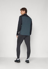 Atletisk outfit bestående af en marineblå jakke med sorte detaljer, tætsiddende sorte bukser og sorte sko. Jakken har en høj krave og lynlås.