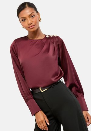 Bluză de satin burgundy cu mâneci lungi și accente cu nasturi pe umăr, asortată cu pantaloni negri și un curea neagră.