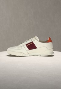 Λευκό low-top αθλητικό παπούτσι με μπορντό πλαϊνή επιφάνεια, χρυσλό λογότυπο «PS Paul Smith» και πορτοκαλί ταμπελάκι στη φτέρνα, εμφανιζόμενο σε ανοιχτή επιφάνεια με γκρι φόντο.