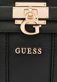 Bolso de mano de cuero negro que presenta un cierre dorado brillante con un logo en forma de G y una placa dorada grabada con "GUESS". Textura suave con detalles cosidos.