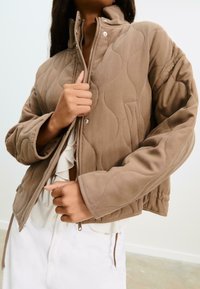 Personne portant une veste matelassée beige sur un chemisier blanc à volants et un pantalon blanc, en zippant le devant de la veste.
