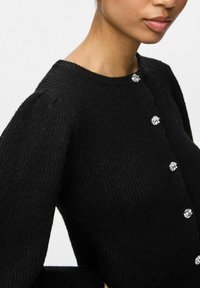 Czarny dziany kardigan z okrągłym dekoltem, teksturowanym materiałem i dekoracyjnymi kryształowymi guzikami z przodu. Bufiaste rękawy dodają objętości.