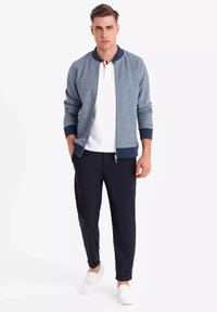 Chaqueta bomber gris con acentos de canalé azul marino, camiseta polo blanca, pantalones oscuros y zapatos blancos sin cordones. Presenta un cierre delantero con cremallera.