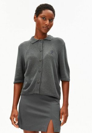 Vrouw met kort haar, gekleed in een grijs vest met knopen en kraag en een grijze rok met een split aan de voorkant, staande tegen een effen witte achtergrond.