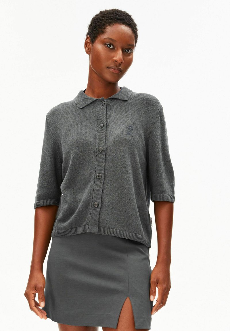 Frau mit kurzen Haaren, die eine graue, geknöpfte Strickjacke mit Kragen und einen grauen Rock mit Schlitz vorne trägt, steht vor einem schlichten weißen Hintergrund.