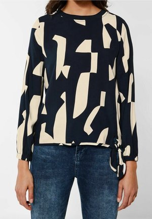 Kvinde iført sort bluse med lange ærmer med beige abstrakte former og sidesløjfe, kombineret med blå denimjeans.