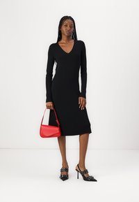 Black ribbed long-sleeve dress with a V-neck, paired with a red handbag and black pointed-toe heels featuring buckle accents.