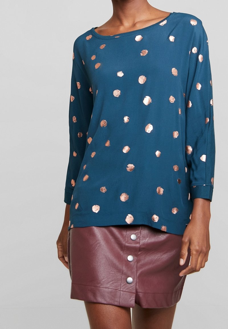Haut à manches longues bleu sarcelle avec pois métalliques couleur or rose associé à une jupe en cuir bordeaux taille haute avec boutons sur le devant.
