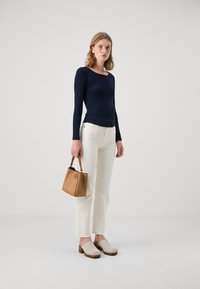 Marineblaues Langarmoberteil mit rundem Ausschnitt, kombiniert mit weißen geraden Jeans. Die Accessoires umfassen eine braune Mini-Handtasche und beigefarbene Clogs.