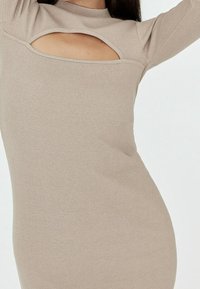 Robe beige côtelée avec un col montant et un décolleté prononcé sur la poitrine, affichant une texture lisse et un design ajusté.