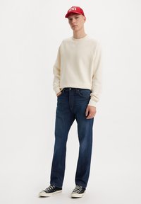 Sudadera beige, jeans azul oscuro y zapatillas negras. La gorra es roja con letras blancas. Corte clásico, estilo casual.
