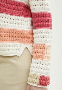 Haut à manches longues crocheté à rayures horizontales rouges, blanches, pêche et beige, porté avec un pantalon beige, montrant un motif en tricot texturé.