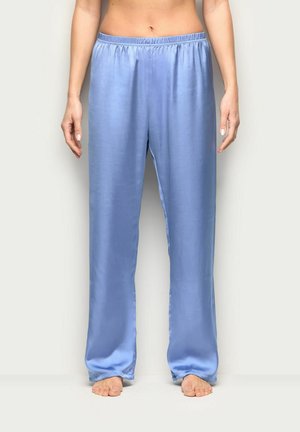 Donna con pantaloni pigiama in satin azzurro chiaro e morbidi, in piedi scalza contro uno sfondo chiaro uniforme, con le mani rilassate lungo i fianchi.