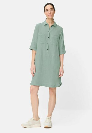 Kvinde stående iført en lysegrøn tekstureret knælange kjole med forlommer og cremefarvede sneakers på hvid baggrund.