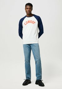 Weißes Langarmhemd mit marineblauen Raglanärmeln und "COWBOY" in Korallenfarbe bedruckt. Kombiniert mit hellblauen Jeans und schwarzen Schnürschuhen.