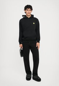 Felpa nera con cappuccio e logo giallo a forma di sorriso, abbinata a pantaloni neri e scarpe nere ricamate. Il modello tiene una borsa nera.
