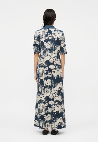 Γυναίκα που στέκεται γυρισμένη με την πλάτη, φοράει μακρύ φόρεμα με ναυτικό μπλε και κρεμ λουλουδάτο σχέδιο, κοντά μανίκια και μαύρα παπούτσια.