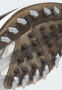 Close-up of a sports shoe sole with black cone-shaped and white arrow-shaped traction studs on a translucent brown base.