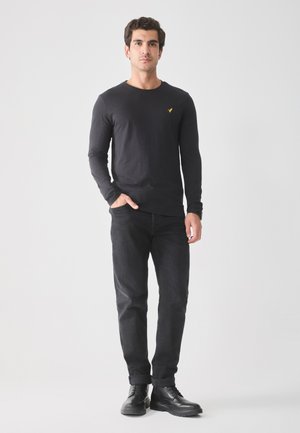 Camisa negra de manga larga con cuello redondo y un pequeño logo amarillo en el pecho, combinada con jeans oscuros y botas negras. Diseño sencillo y ajustado.