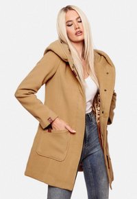 Tan huva jacka med framknappar, sidofickor och en mjuk tygtextur, bärs över en vit topp och kombineras med denimjeans.