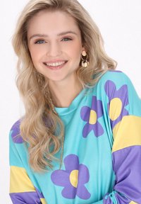 Camisa de manga larga en turquesa con patrones florales en amarillo y púrpura. El modelo lleva pendientes de oro y tiene el cabello rubio y ondulado.