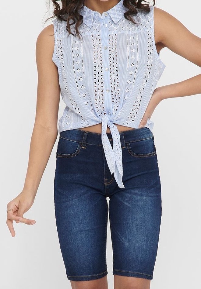 Blouse bleu clair sans manches avec des détails en broderie anglaise et un nœud à l'avant, associée à un short en denim foncé arrivant à mi-cuisse.