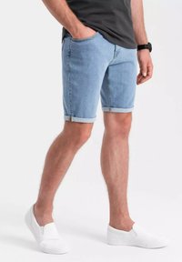 Pantalones cortos de mezclilla para hombre en azul claro, con dobladillo enrollado, bolsillos laterales y passadores para cinturón, combinados con zapatos blancos de tipo slip-on.