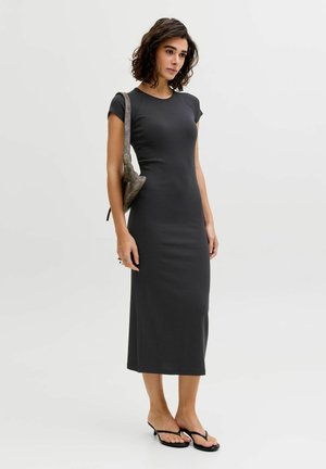 Mujer con cabello rizado oscuro hasta los hombros, vestida con un vestido midi negro ajustado de manga corta, llevando un bolso gris al hombro y sandalias negras.