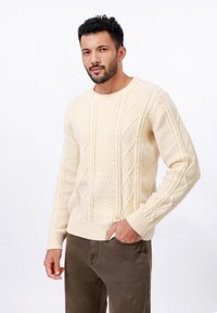 Pull en maille torsadée crème avec un col rond, orné de motifs en losange et de poignets côtelés, associé à un pantalon marron foncé.