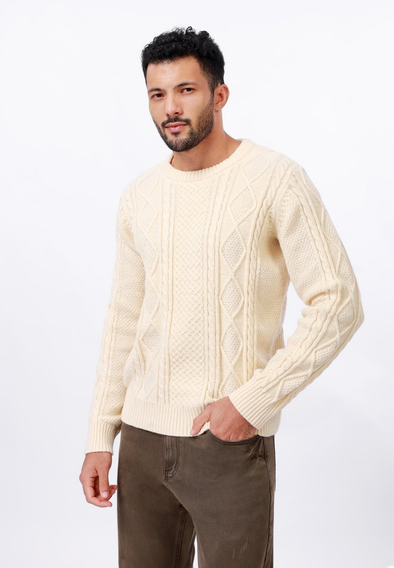 Pull en maille torsadée crème avec un col rond, orné de motifs en losange et de poignets côtelés, associé à un pantalon marron foncé.
