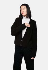 Schwarzer, strukturierter Cardigan mit tiefem V-Ausschnitt, lockerer Passform und zwei schwarzen Knöpfen auf der Vorderseite, kombiniert mit einem weißen Rollkragenpullover darunter.